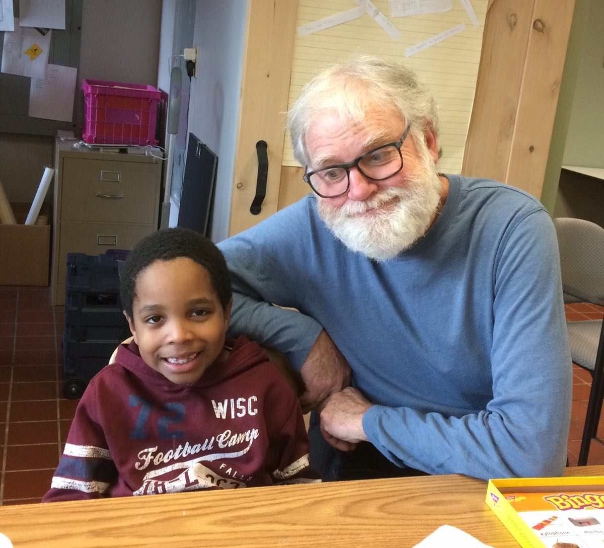 A young boy and an older man sitting together at a table, both smiling. The boy is wearing a maroon hoodie with football text, and the man has white hair and a beard, wearing glasses and a blue long-sleeve shirt. They are indoors, in a room with a wooden wall and various items in the background.