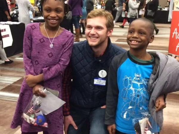 A young man is with two children at an indoor event or fair, smiling for the photo. The girl on the left is wearing a purple dress, and the boy on the right is wearing a blue graphic t-shirt and holding a small bag.