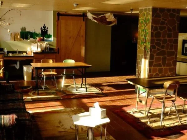 Interior of a cozy cafe or restaurant with wooden furniture, colorful rugs, and a stone accent wall. There are tables and chairs arranged throughout, with some tables set near a small counter or bar area. Sunlight casts shadows across the hardwood floor.