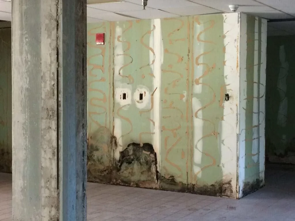 Interior photo of a damaged wall with exposed electrical outlets and graffiti-like orange markings, showing signs of water damage at the bottom.
