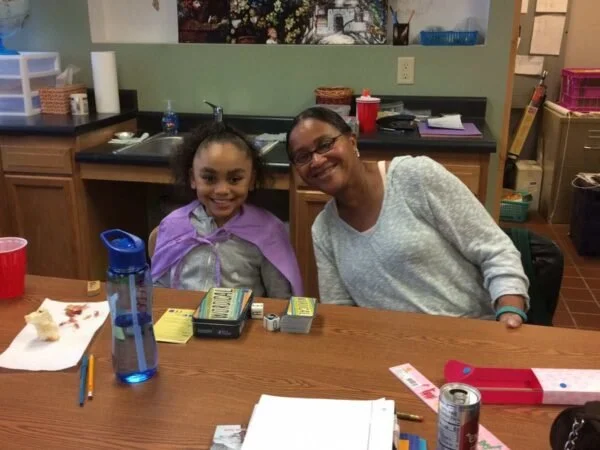 A woman and a young girl sitting at a table in a kitchen, smiling at the camera. The table has various items including a water bottle, a red cup, a can, and some papers.