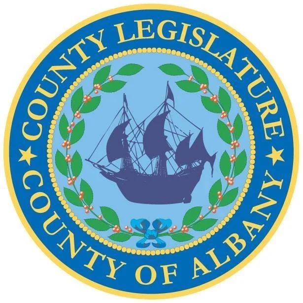 Seal of Albany County Legislature featuring a sailing ship, green laurel branches with berries, and a blue ribbon at the bottom.
