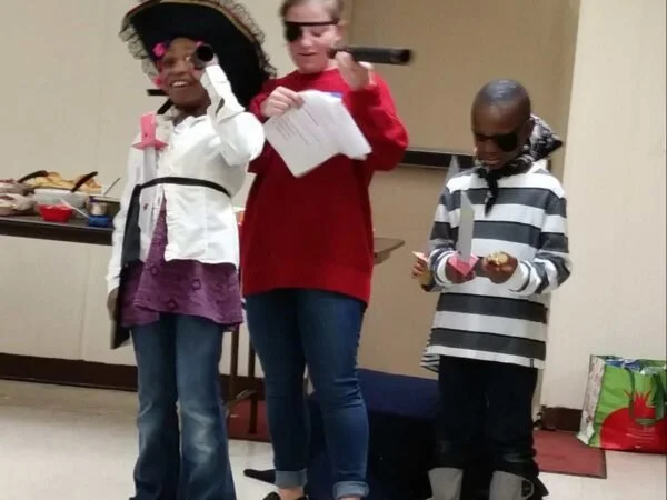 Three children dressed in costumes, with one holding a microphone, standing in a room near a table with food and a colorful gift bag.
