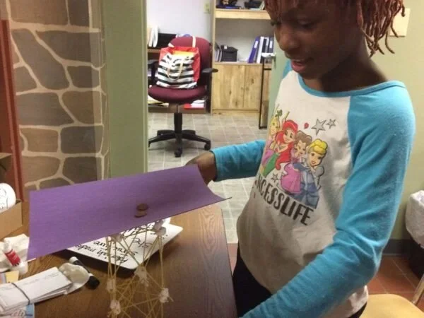 A young girl with dreadlocks wearing a princess-themed shirt, looking at a small paper diorama with a paper boat and other craft materials on a wooden table.