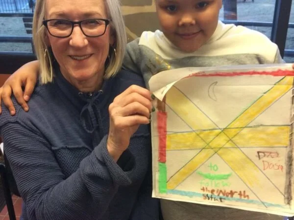 An older woman with glasses and shoulder-length blonde hair smiling, sitting next to a young boy holding a colorful school project sign. The woman has her arm around the boy, and they are indoors.