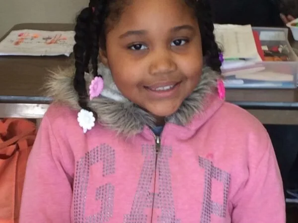 A smiling young girl with braided hair decorated with pink and white flower hair clips, wearing a pink jacket with a fur-lined hood, sitting at a desk with books and papers in the background.