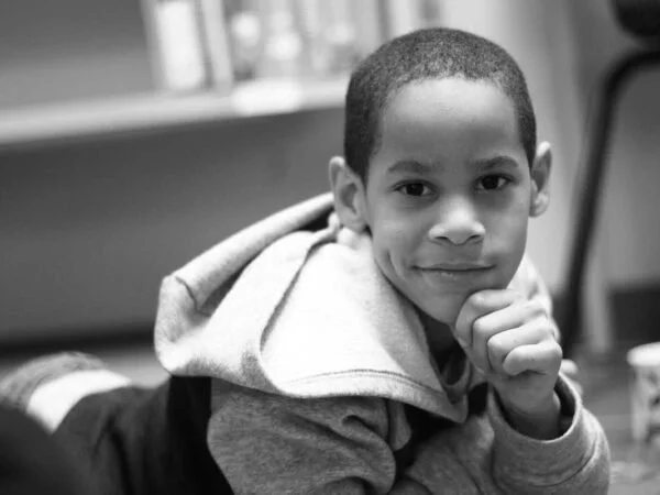 A young boy with short hair in a hoodie, resting his chin on his hand, sitting indoors in a room with windows.