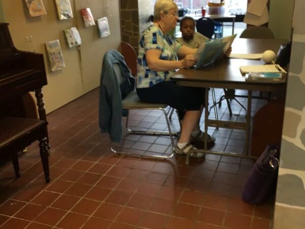 An elderly woman and young girl sitting at a table in a cozy cafe or library, looking at a newspaper together.