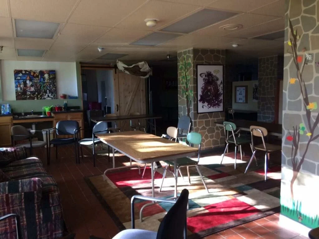 Interior of a room with a wooden table, multiple chairs, a sofa with a plaid pattern, and wall decorations including paintings and tree mural, with sunlight streaming through windows.