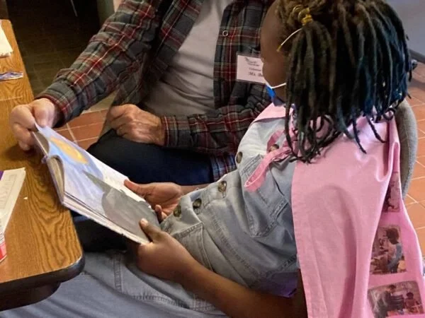 A young girl with braided hair and a pink backpack sitting at a wooden table, reading a book with an adult.