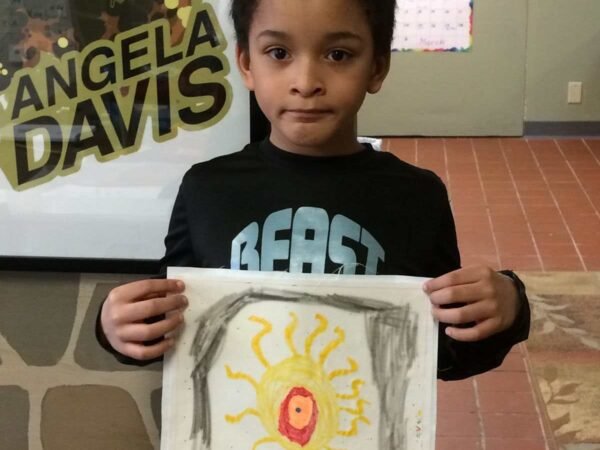 A young boy holding a drawing of a yellow sun with orange and red details, standing in a room with a tiled floor and a display board with the words 'Angela Davis' in the background.