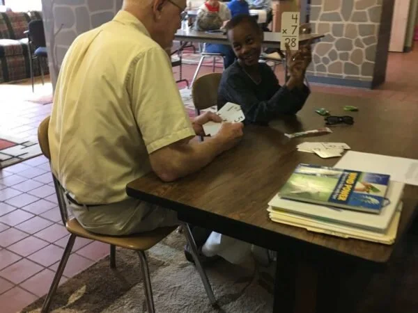 An older man and young girl sitting at a table playing a card game inside a classroom or library. The girl is smiling and holding up a card.
