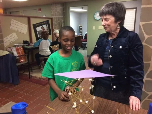 A young boy in a green shirt shows a paper and a structure made of sticks and marshmallows to an older woman in a classroom or community space.