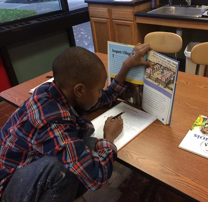 A young boy with a shaved head wearing a plaid shirt reads a book at a table in a library or classroom.