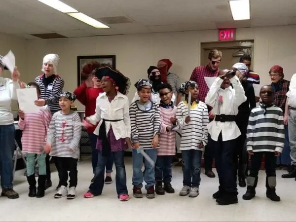 Children dressed as pirates and captains, standing in a line indoors, with adults around them, some dressed in costumes and hats, in a decorated room.