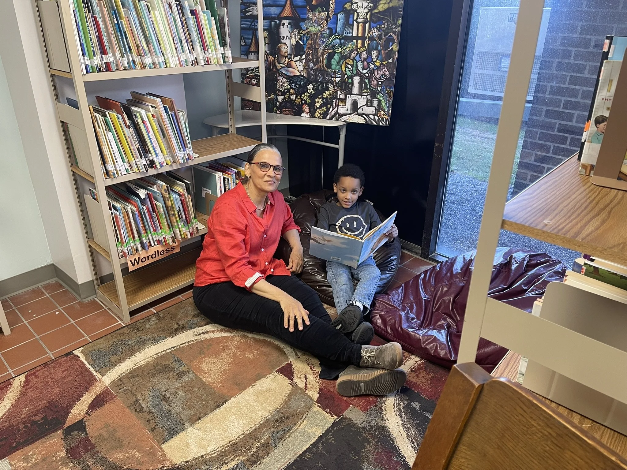 A woman and a young boy sitting on the floor of a library or bookstore, reading a book together. There are bookshelves behind them, a colorful stained-glass window in the background, and a patterned rug on the floor.