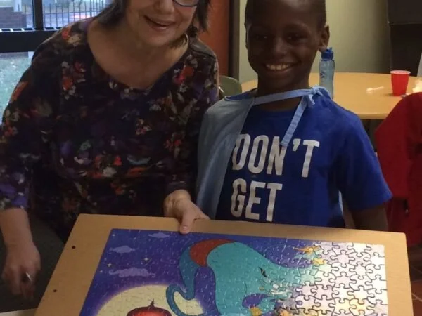A woman and a young boy smiling and working on a completed colorful puzzle of a unicorn with a rainbow mane.