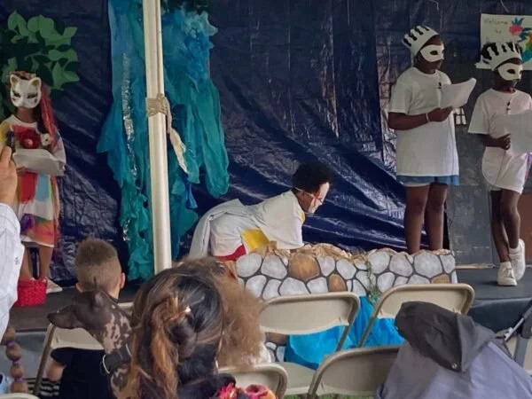 Children performing a play on stage, with some wearing animal masks and costumes, and an audience watching.