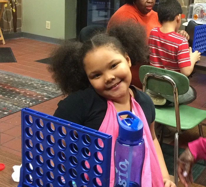 A smiling young girl with curly hair wearing a black shirt and pink apron, sitting at a table in a classroom setting, with a blue water bottle in front of her and other children in the background.