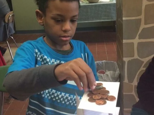 A young boy wearing a blue shirt and gray sleeves holds pennies and places them on a white paper stack.