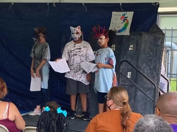 Three children wearing animal masks and holding papers stand on a small stage with a blue backdrop, performing in front of an audience.