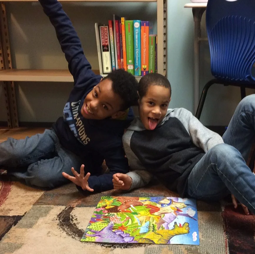 Two children lying on a rug, excited and smiling, with a colorful jigsaw puzzle of cartoon dinosaurs and scenery on the floor.