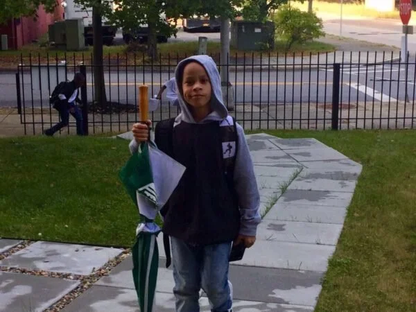A young boy standing on a sidewalk, holding a green and white umbrella and wearing a backpack, with a school building and another person walking in the background.