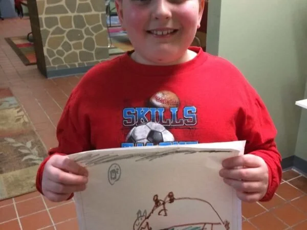 A smiling young boy in a red shirt holding a piece of paper with a child's drawing, standing indoors.