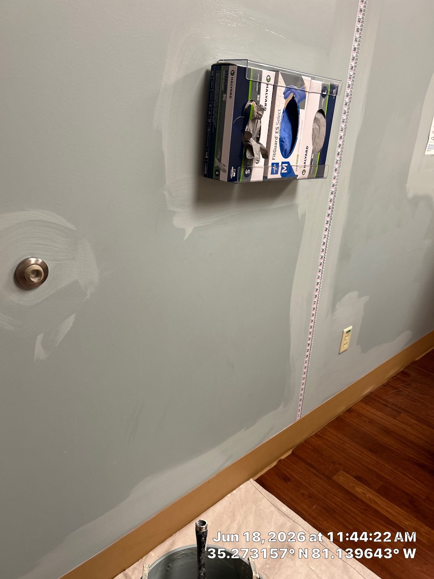 Wall partially painted with light color, with a measuring tape or ruler stretched across it. There is a box of a hoverboard and a paint roller tray on the floor. Date, time, and coordinates are overlaid on the image.