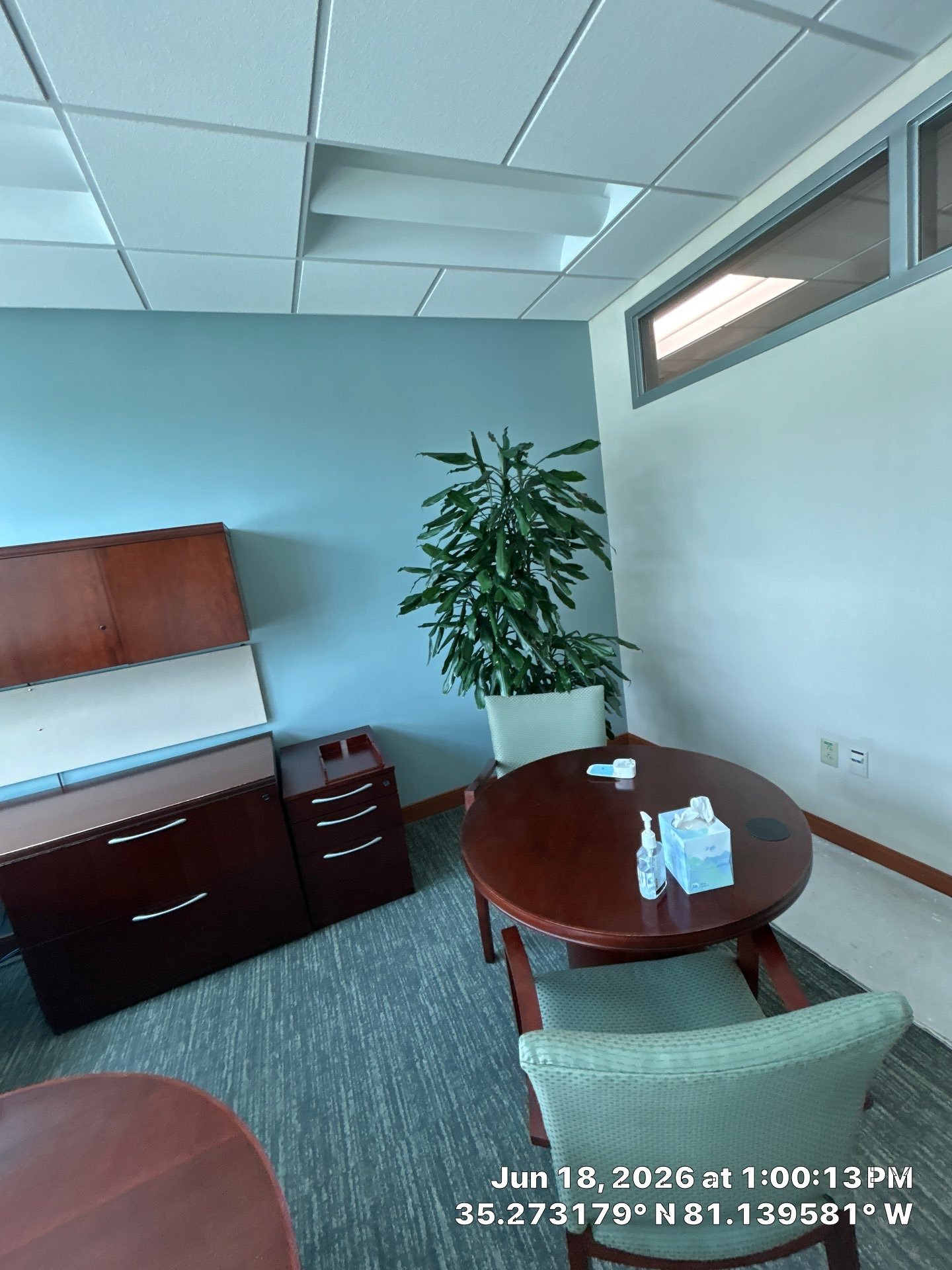 Office room with a round wooden table, upholstered chairs, a tall green plant, wall-mounted cabinets, tissue box, hand sanitizer, and a window near the ceiling. The photo is timestamped June 18, 2026, 1:00:13 PM, with GPS coordinates 35.273179°N 81.1