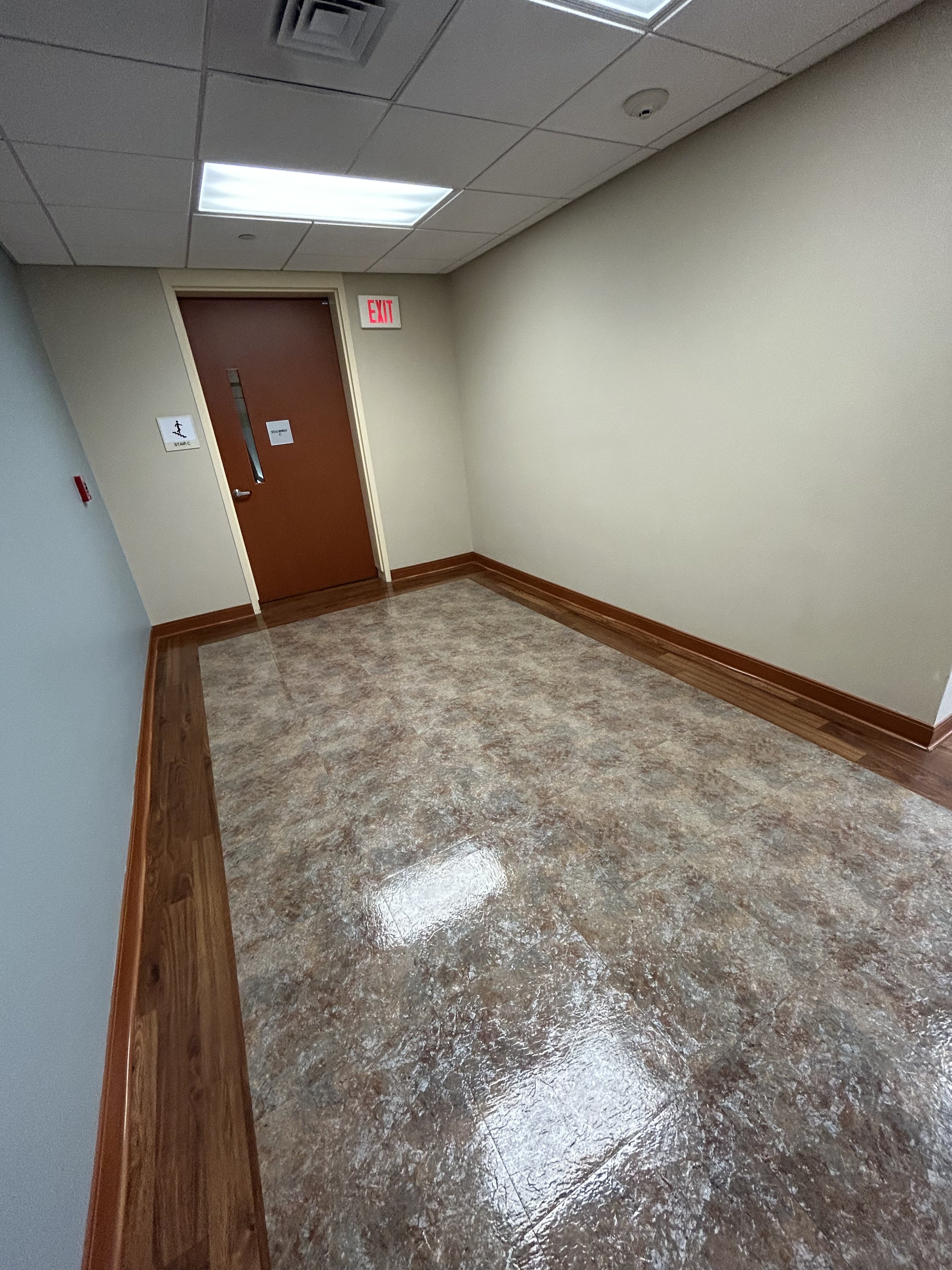 Empty hallway or corridor with beige walls, brown door with a silver handle, and a red EXIT sign above the door. The floor is a polished, brownish tile, and the ceiling has square tiles with one fluorescent light and one smoke detector.