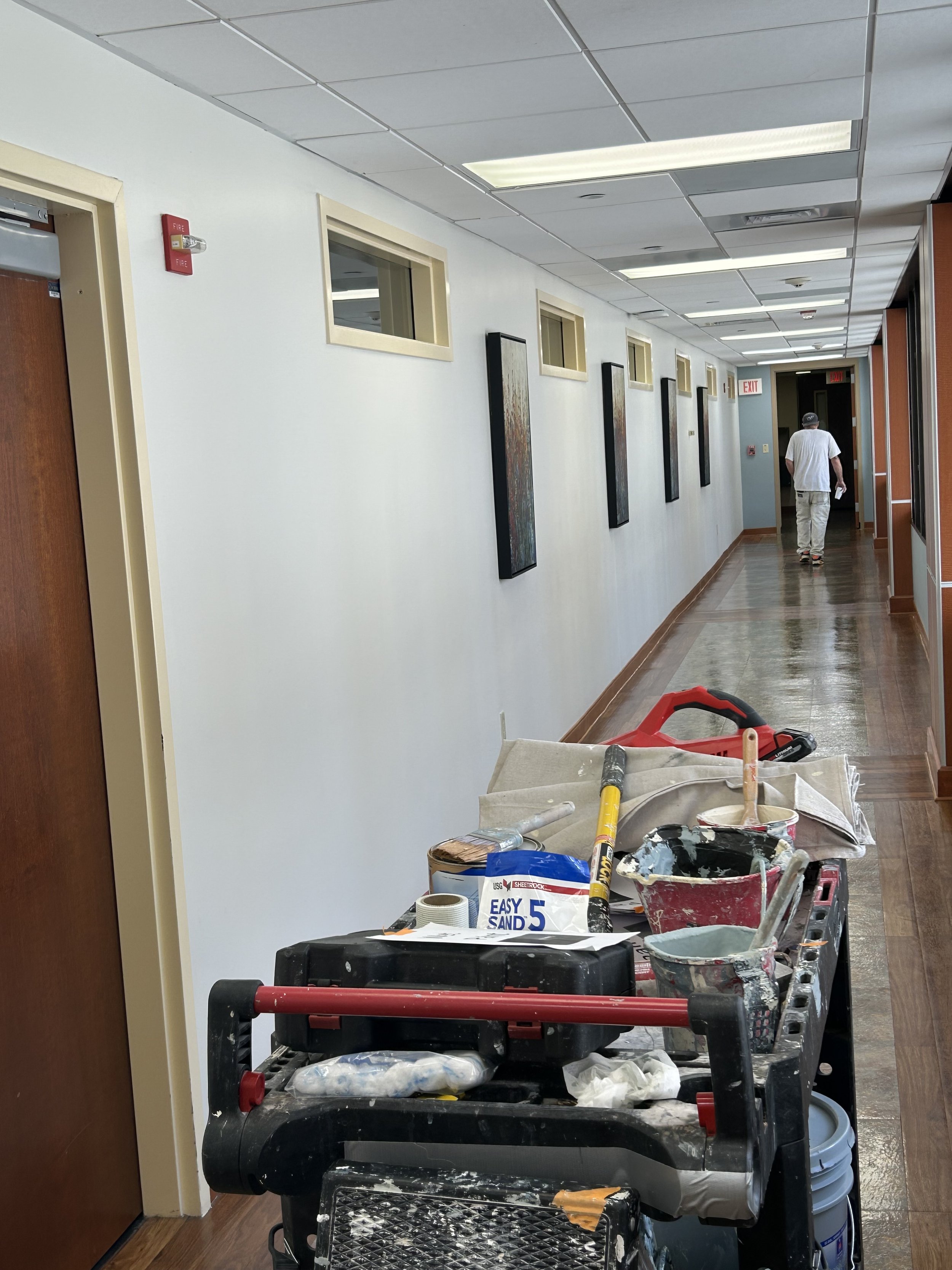 A hallway under renovation with painting supplies and tools on a rolling cart in the foreground, a man walking away in the distance, and several small windows lining the white wall.