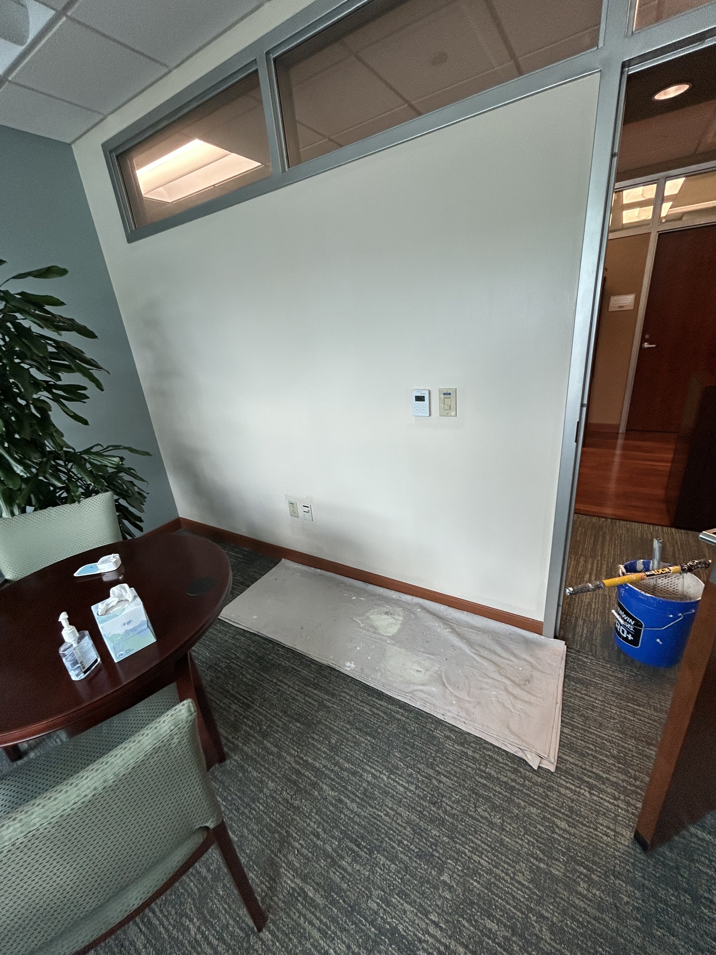 An office corner with a white wall, a window near the ceiling, a potted plant, a brown table with hand sanitizer and tissues, a carpeted floor, a section of a patterned armchair, and a room under renovation with a blue bucket and tools.
