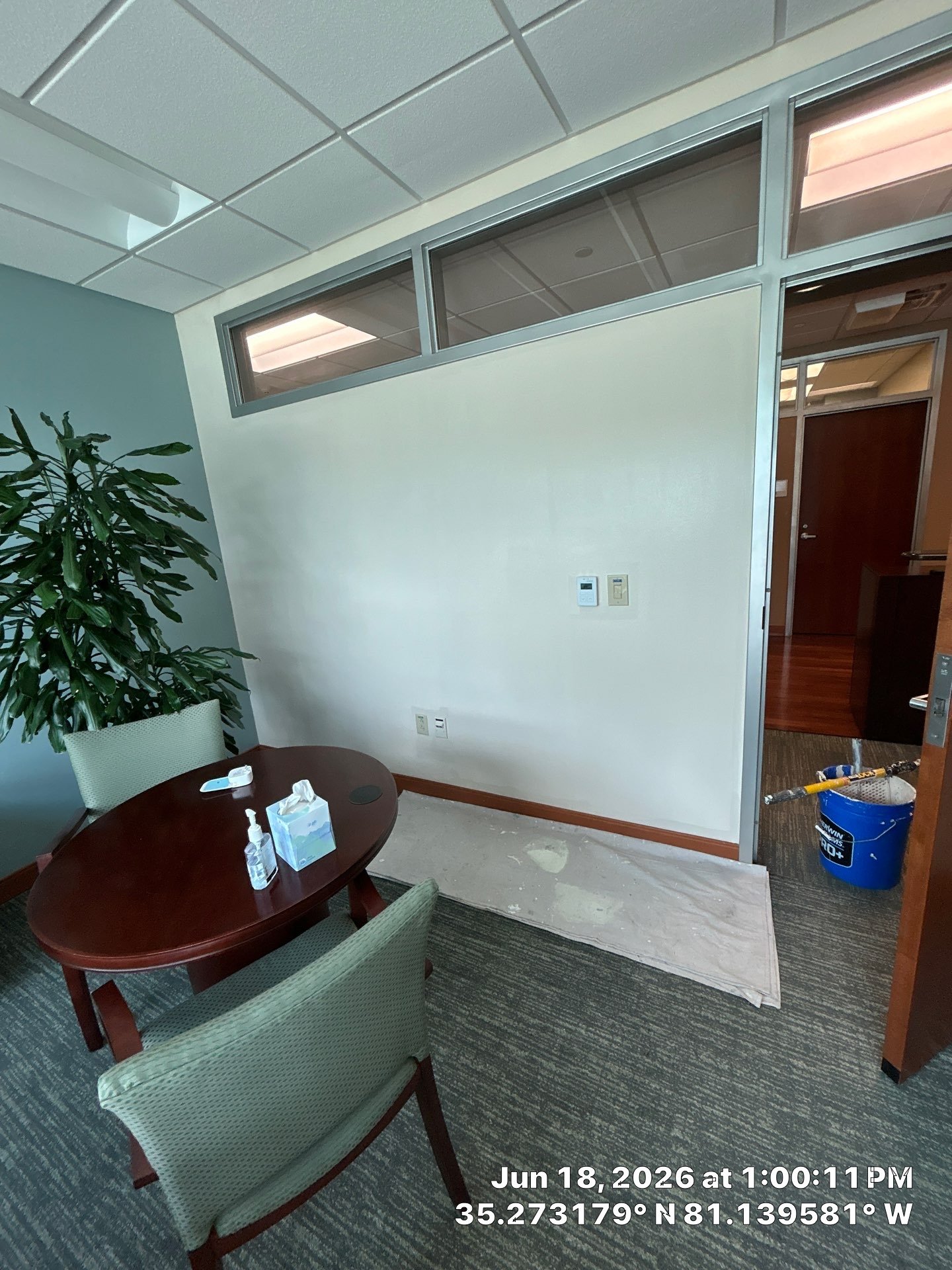 Office room with a table, chairs, plant, and partially uncovered carpet, with construction supplies and a partially covered floor. A windowed wall separates this room from an adjoining space. The room has a white wall with visible switches and outlet