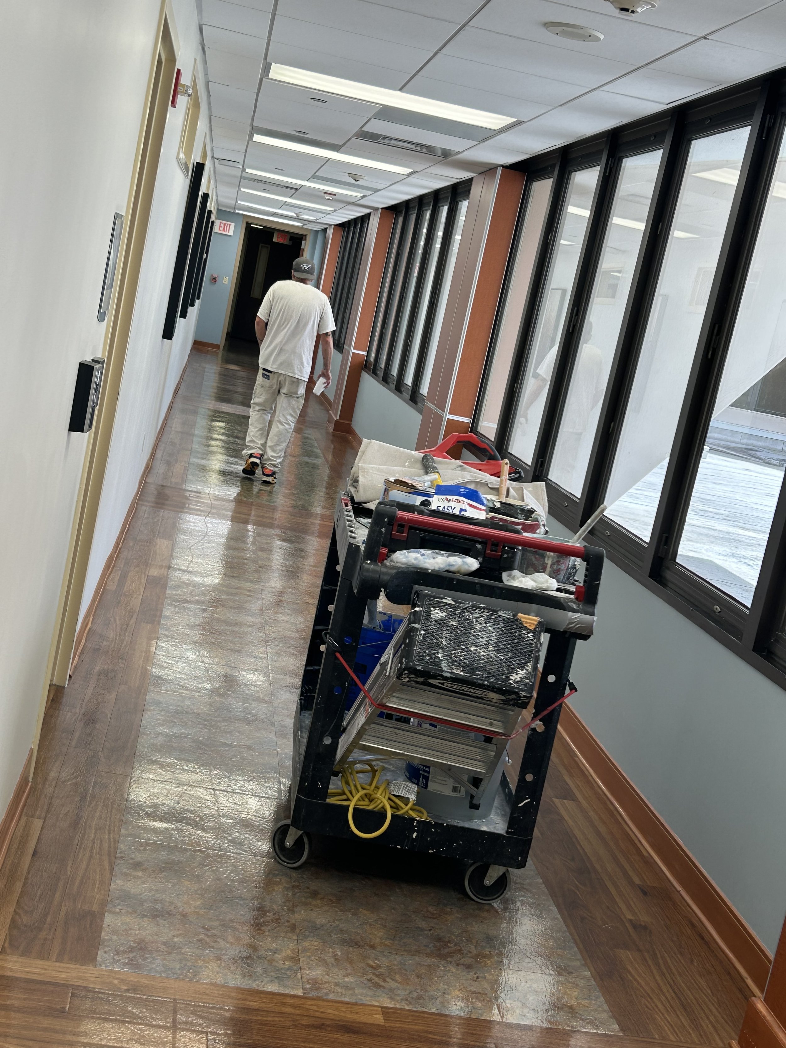 A man walking down a hallway with a construction cart filled with tools and supplies. The hallway has windows on one side with a blue wall on the other. The ceiling has fluorescent lights, some of which are open or damaged. The floor is wet, likely f