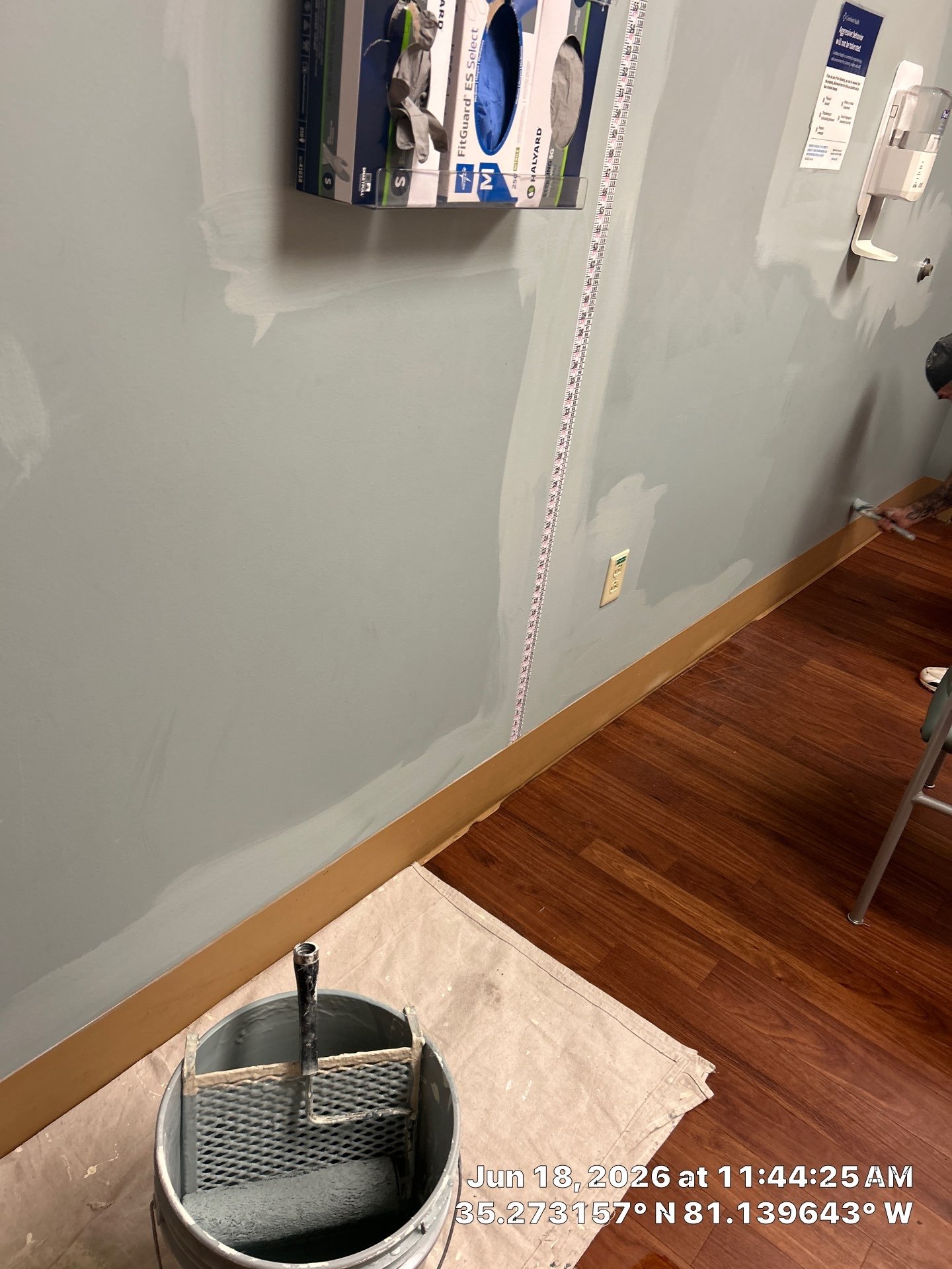 Interior wall in the process of painting or renovation, with patches of primer or paint, a tape measure on the wall for measurement, a bucket with tools in the foreground, and a temperature and GPS coordinate overlay at the bottom right corner. The d