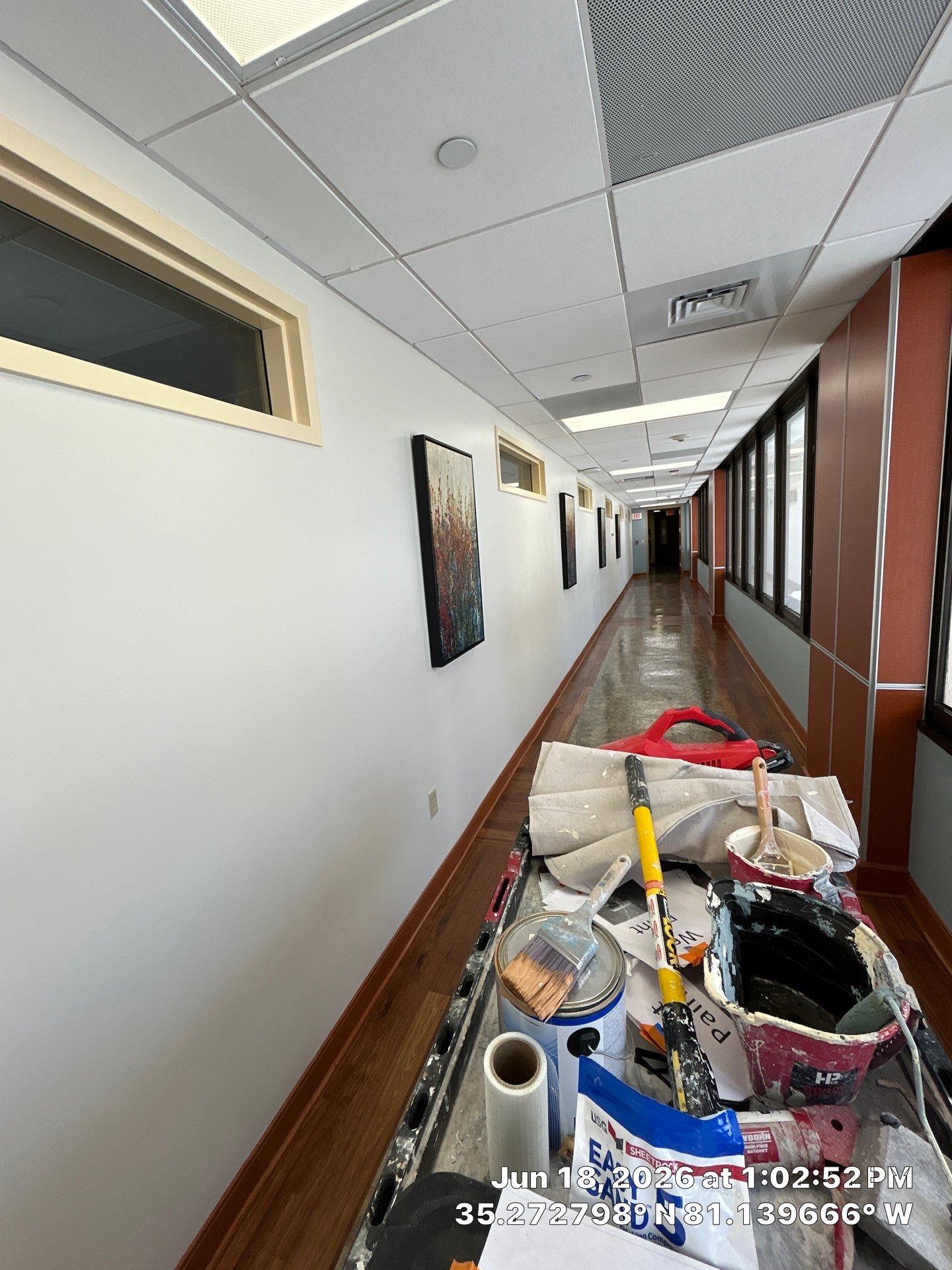 A long hallway with white walls, wooden baseboards, framed artwork, and large windows along the right wall. The ceiling has white tiles with some ventilation grilles and light fixtures. On a table in the foreground are painting tools, paint cans, bru