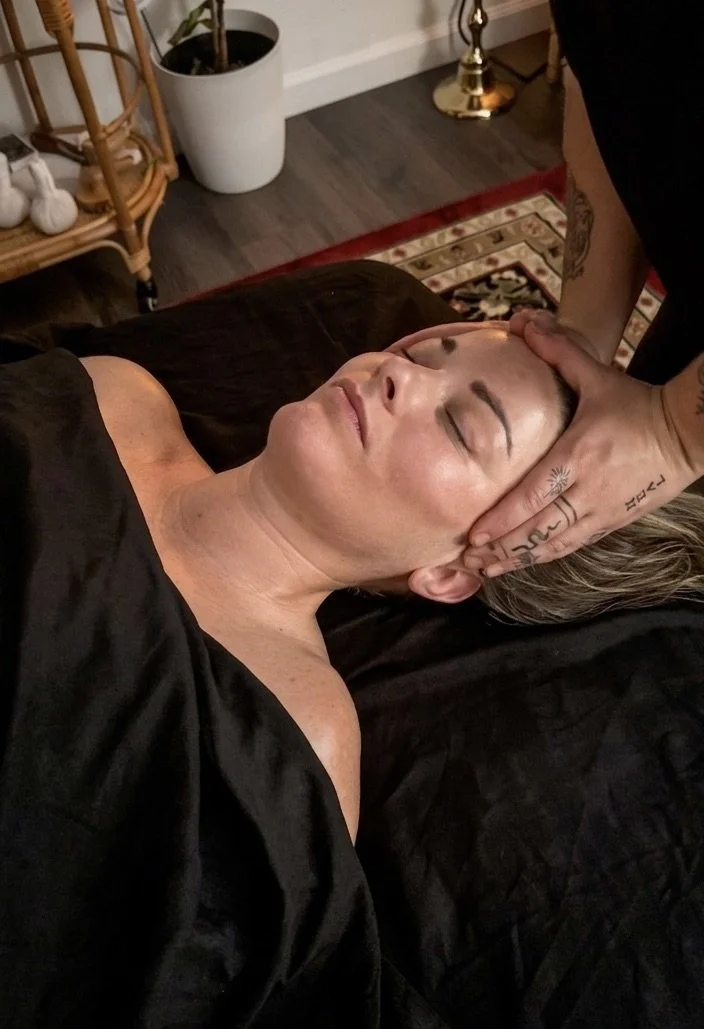 A woman receiving a TMJ face massage treatment from a therapist whose hands are on her forehead. The woman has her eyes closed and appears relaxed.