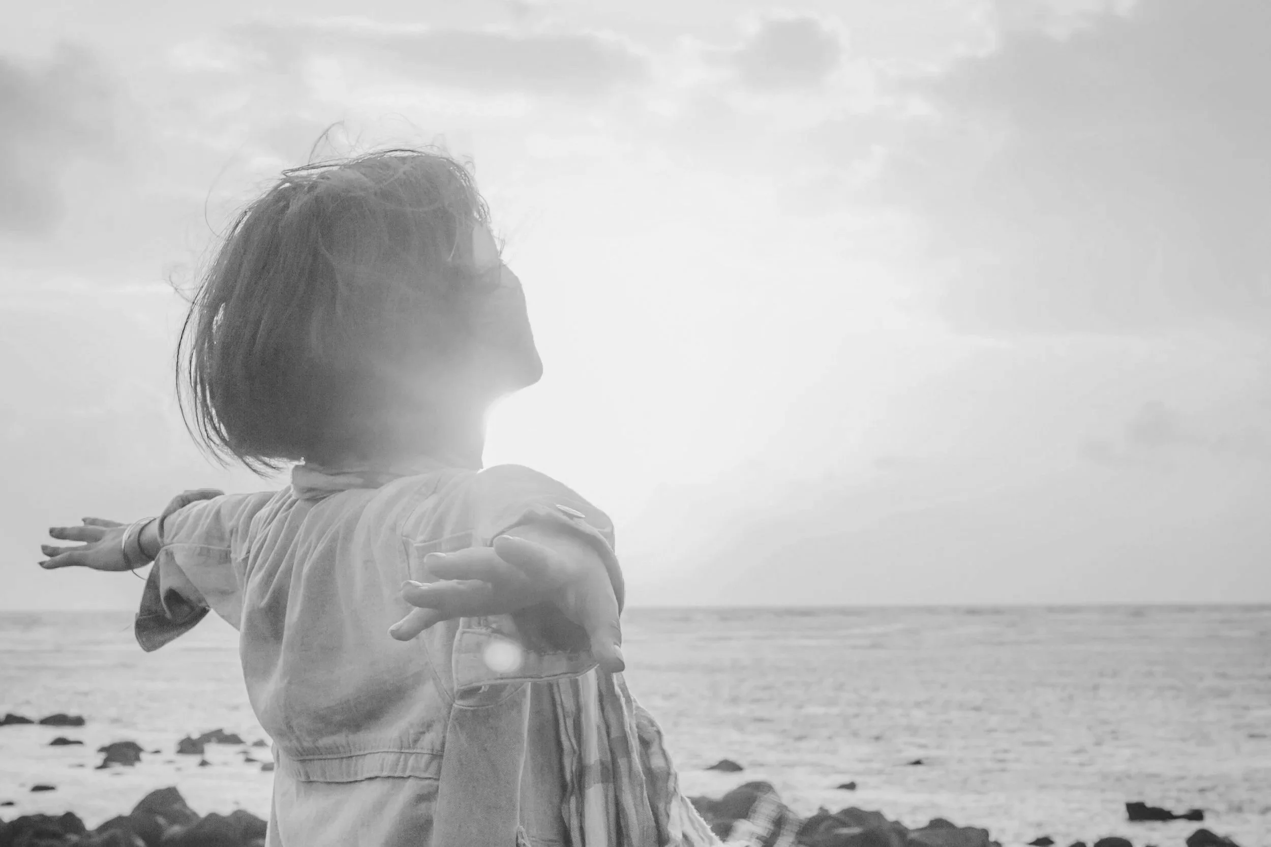 A person with shoulder-length hair is hugging another person at the beach, both facing toward the ocean, with arms outstretched. The photo is in black and white, showing the cloudy sky and shoreline in the background.