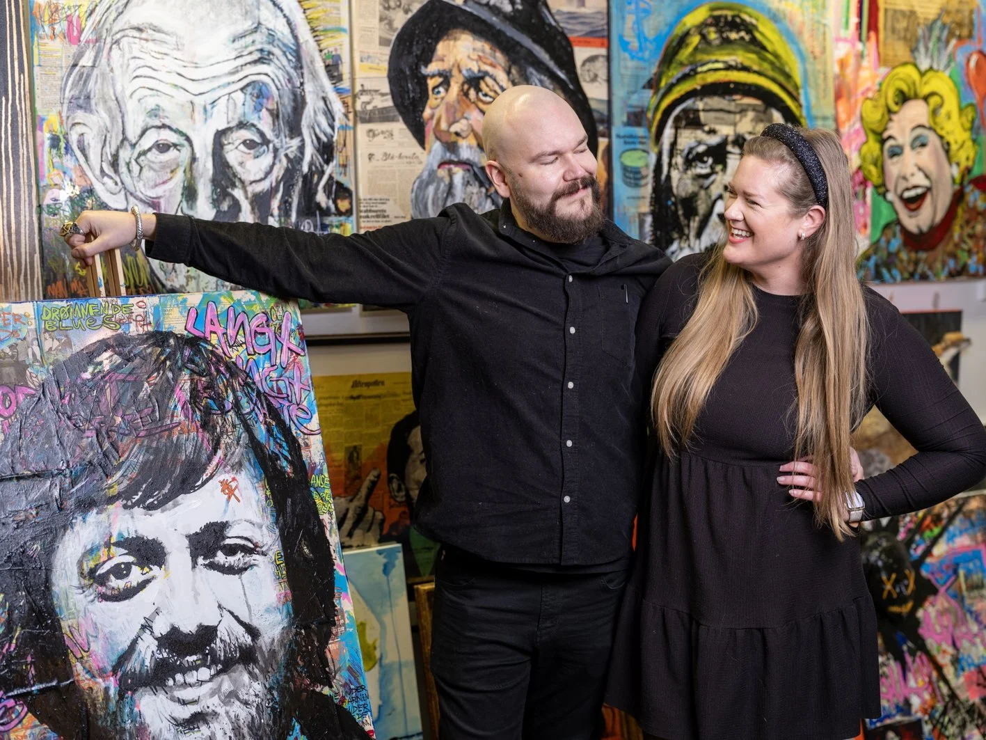 To personer som smiler og står foran en vegg med flere kunstverk, inkludert portretter av kjente personer, i en kunstgalleri.