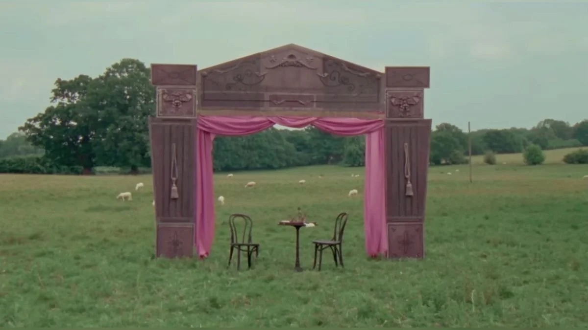 A decorative wooden outdoor wedding arch with pink curtains, a small round table with two chairs in front of it, set in a grassy field with sheep grazing and trees in the background.