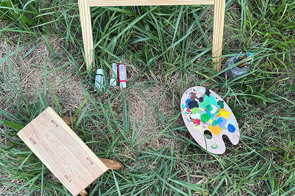 Outdoor painting setup on grassy ground with a wooden easel, a blank wooden board, a paint palette with various colors, and paint bottles.