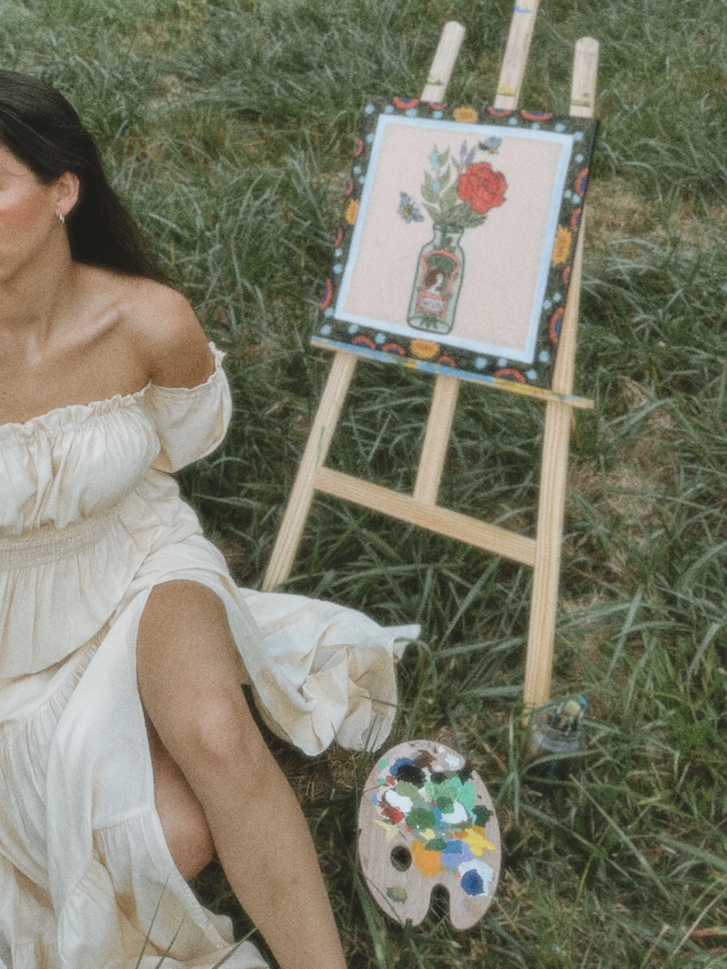 A woman in a white dress lying on the grass next to an easel with a painting of a bottle and a red flower in a vase, surrounded by green grass and trees.