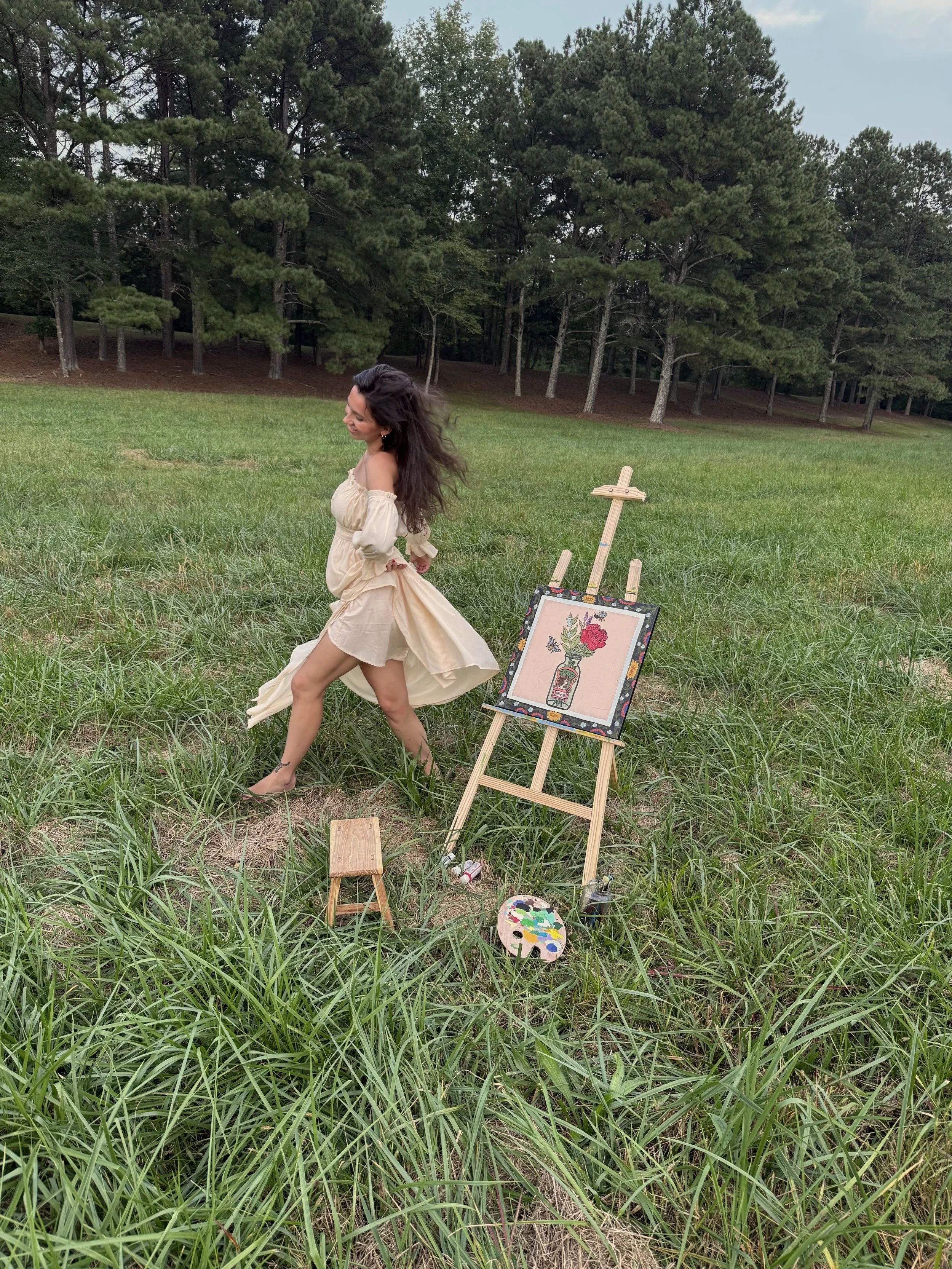 A woman in a beige dress dancing or twirling in a grassy field with an artist's easel and painting supplies nearby, surrounded by tall grass and trees in the background.