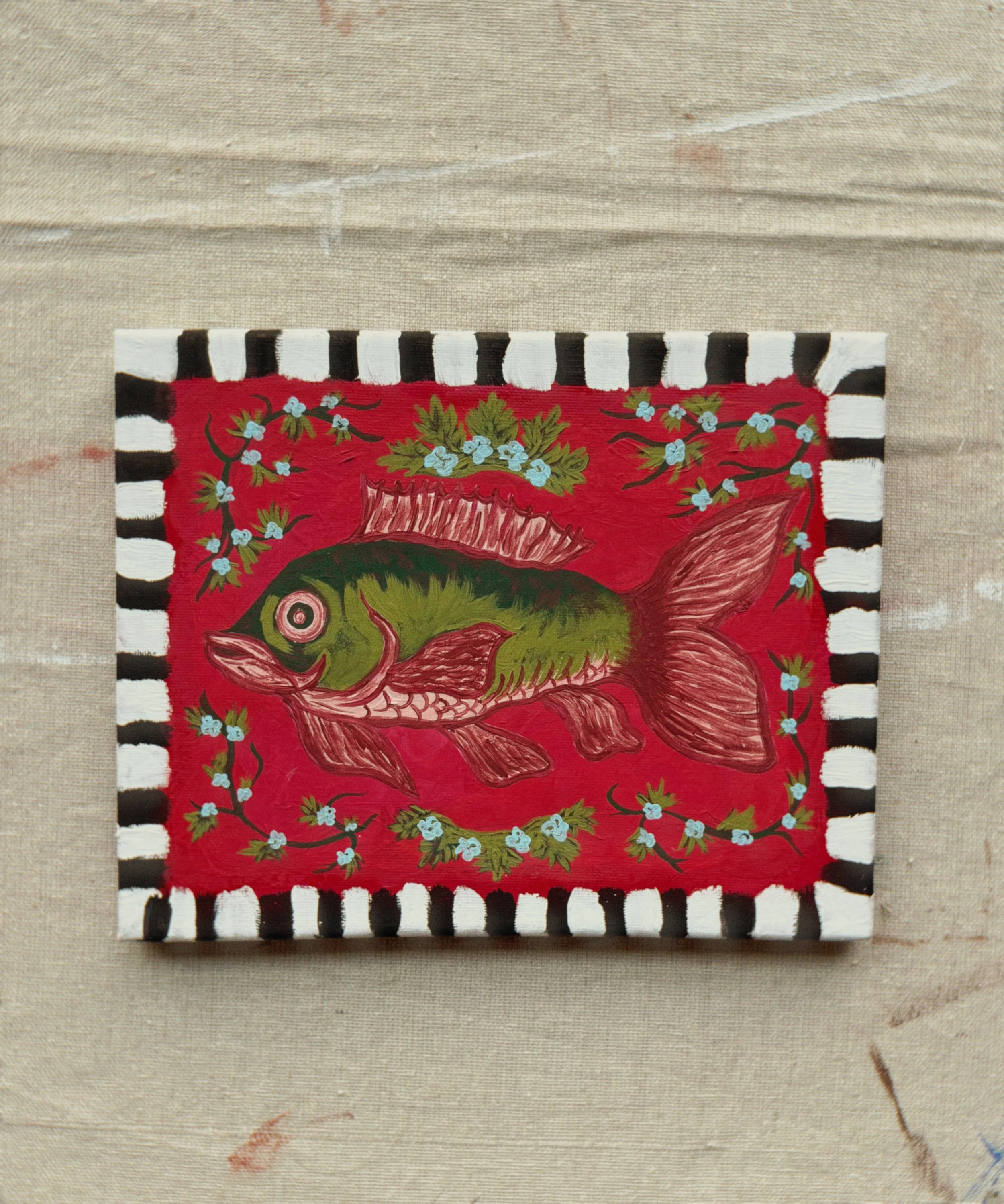 A close-up of a colorful, hand-painted rectangular artwork featuring a fish, surrounded by decorative floral elements and enclosed by a black and white striped border, laid on a neutral fabric background.