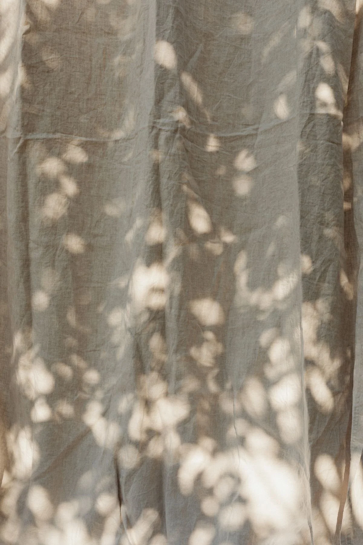 Close-up of a sheer, light-colored curtain with sunlight shining through, creating a pattern of bright spots and shadows.