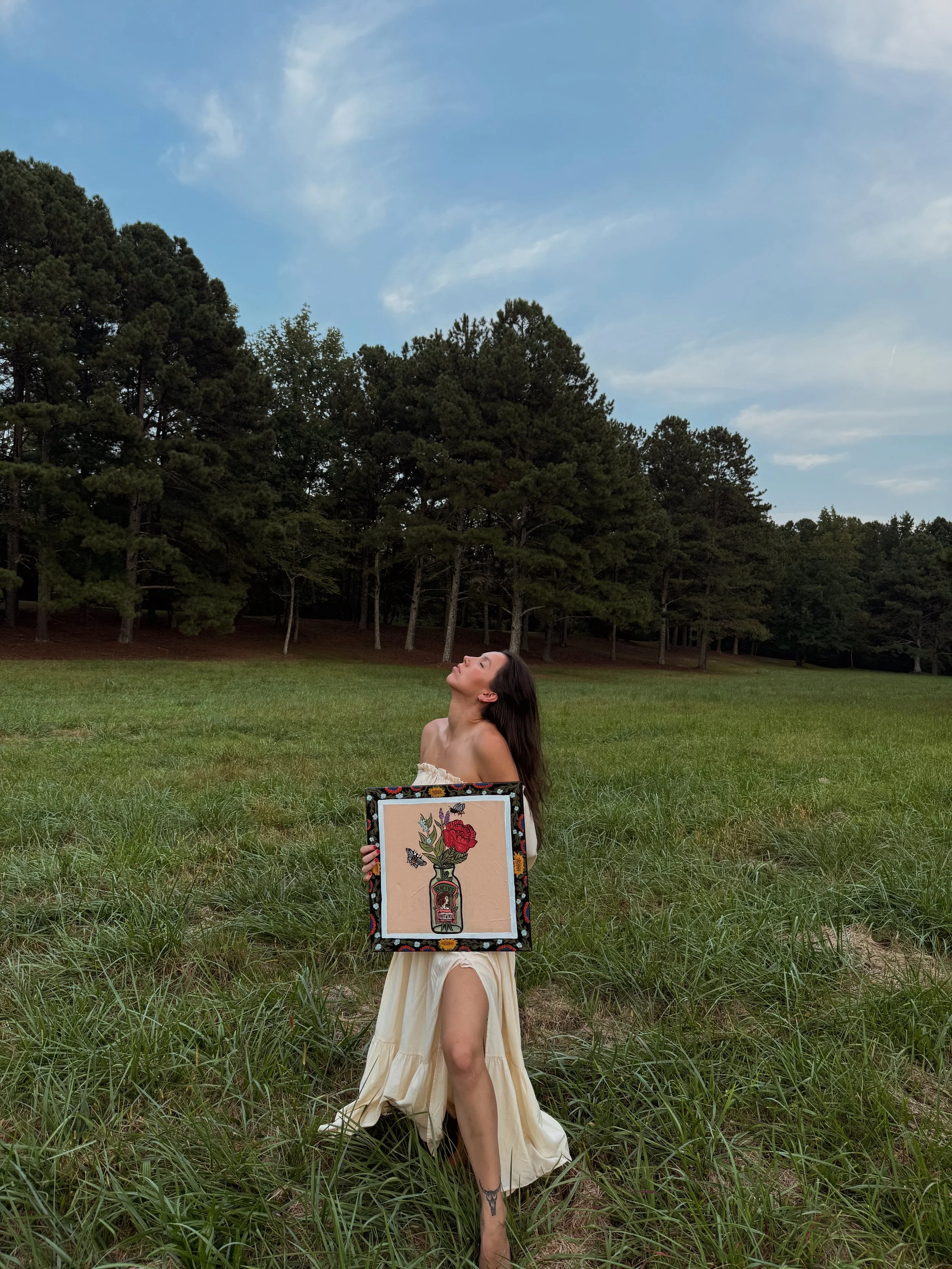A woman with long dark hair, standing in a grassy field, holding a framed artwork of a vase with flowers and butterflies. She is wearing a cream-colored dress with a slit, and the background features a line of tall trees and a partly cloudy sky.