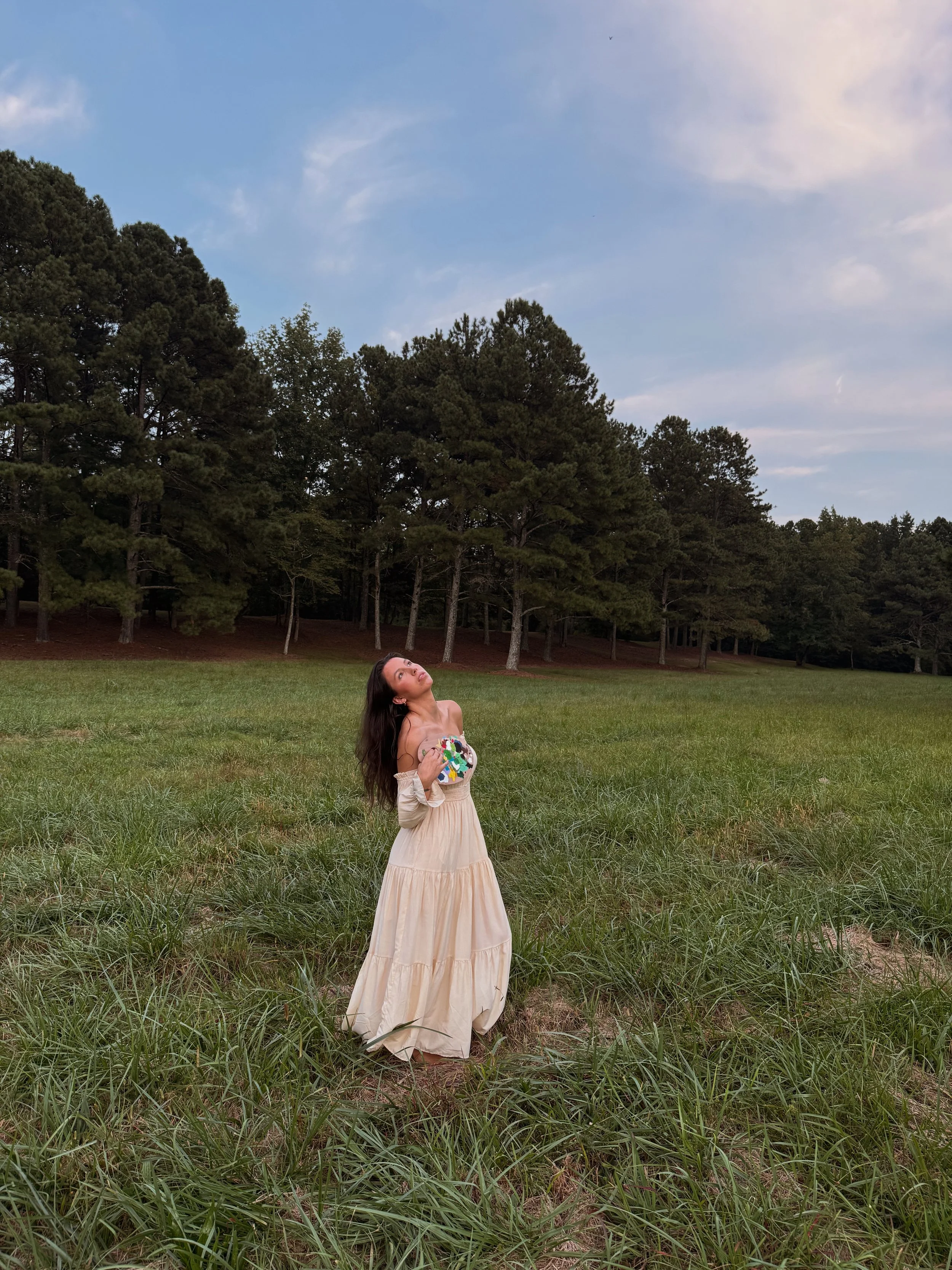 A woman in a flowing cream-colored dress standing in a grassy field, holding a colorful object, with tall trees and a partly cloudy sky in the background.
