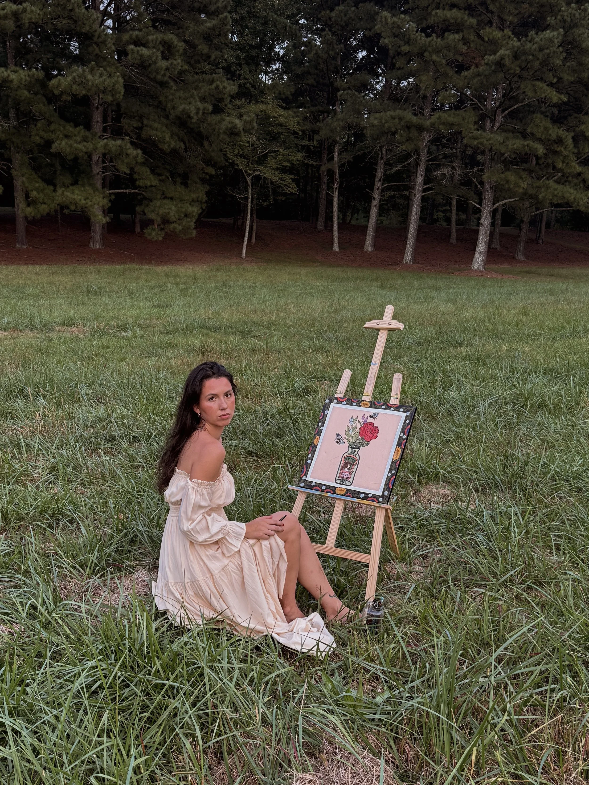 A woman with dark hair and light skin, wearing an off-shoulder cream-colored dress, sitting in a grassy field, looking at the camera. Behind her, there is a painting of a flower in a vase on an easel and a small bottle on the grass. Trees with green foliage are in the background.