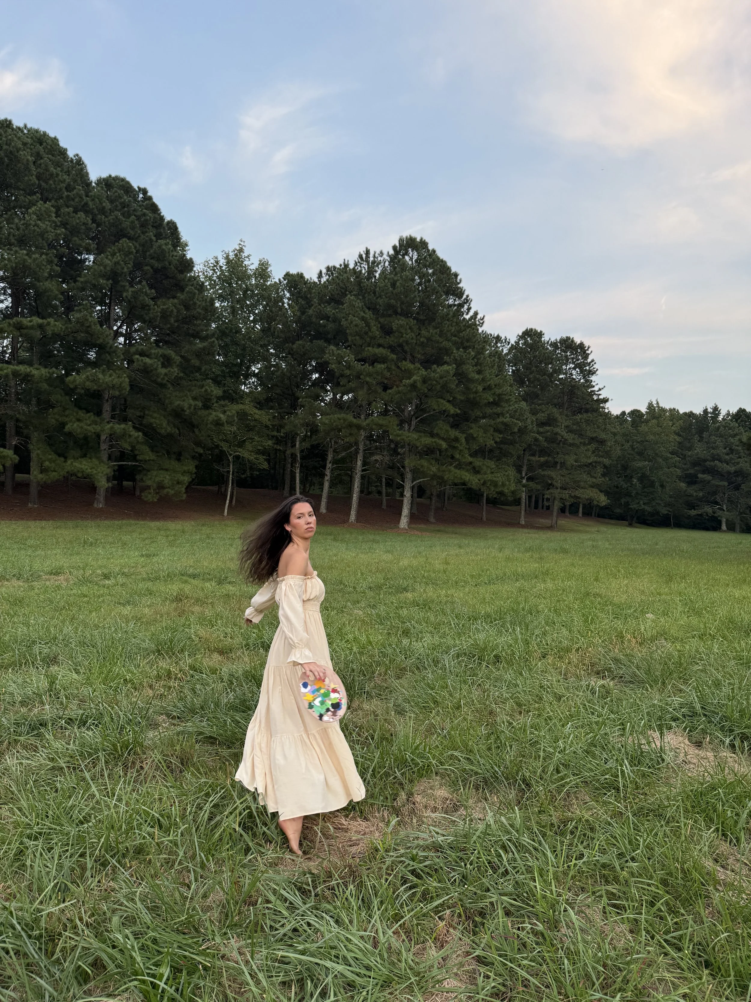 Woman in a cream dress walking barefoot through tall grass in a field, holding colorful floral paper cutouts, with a backdrop of dense trees and a partly cloudy sky.